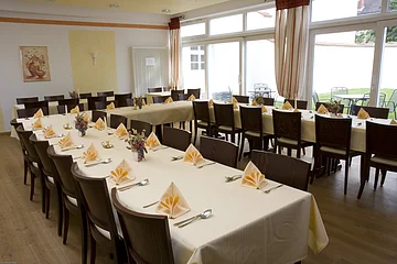 Ein großer Raum mit gedeckten Tischen, Stühlen und gefalteten Servietten, Fenster im Hintergrund.