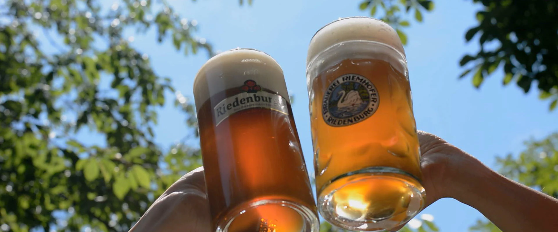 Zwei Personen mit Bierkrüge in der Hand stoßen im Sonnenschein an.