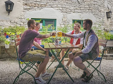 Vier Personen sitzen an einem Tisch im Freien und stoßen mit Bierkrügen an. Im Hintergrund sind Pflanzen und eine Steinmauer.
