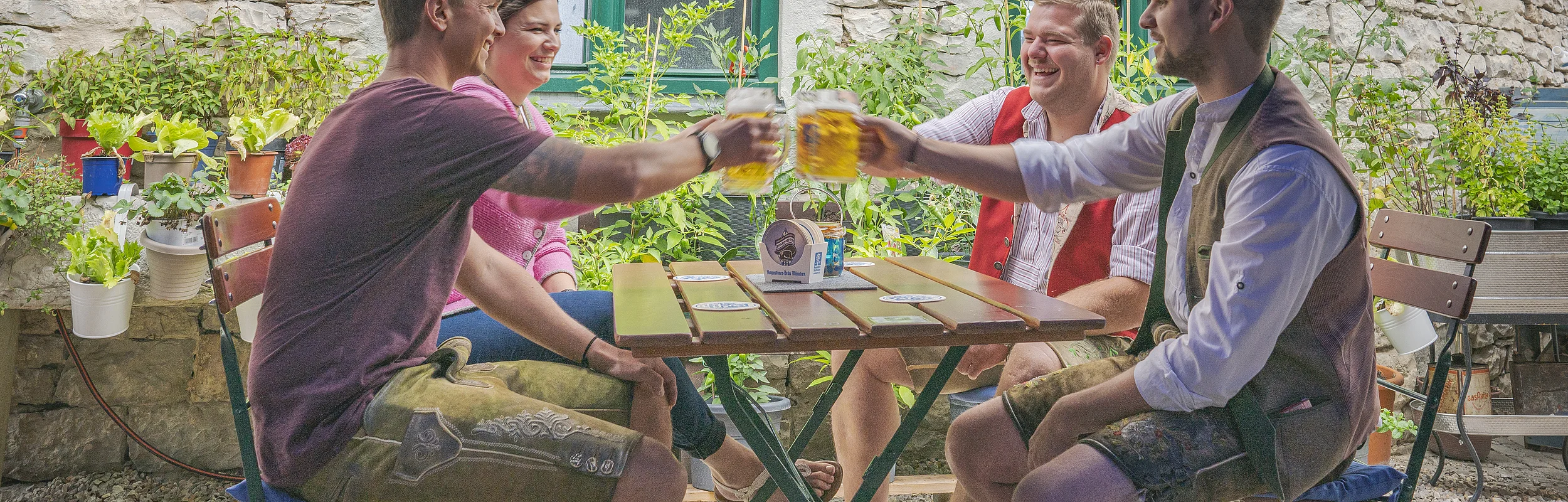 Vier Personen sitzen an einem Tisch im Freien und stoßen mit Bierkrügen an. Im Hintergrund sind Pflanzen und eine Steinmauer.