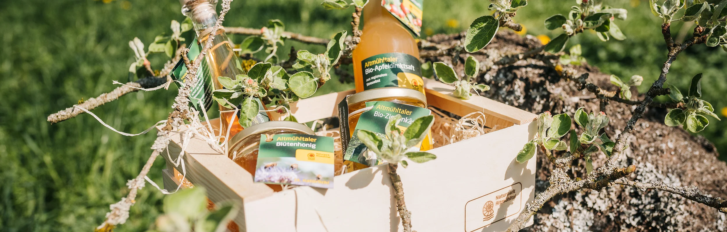 Holzkiste mit Bio-Produkten, darunter Apfelsaft, Honig und Likör, auf einem Baumstamm im Freien platziert.