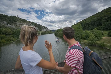 Eine Frau mit weißen Shirt und ein Mann mit Rucksack stehen mit dem Blick auf den Main-Donau-Kanal gerichtet auf der Holzbrücke am Geländer. Der Himmel ist sehr bedeckt. Die beiden Ufer erstrahlen im dunklen grün.