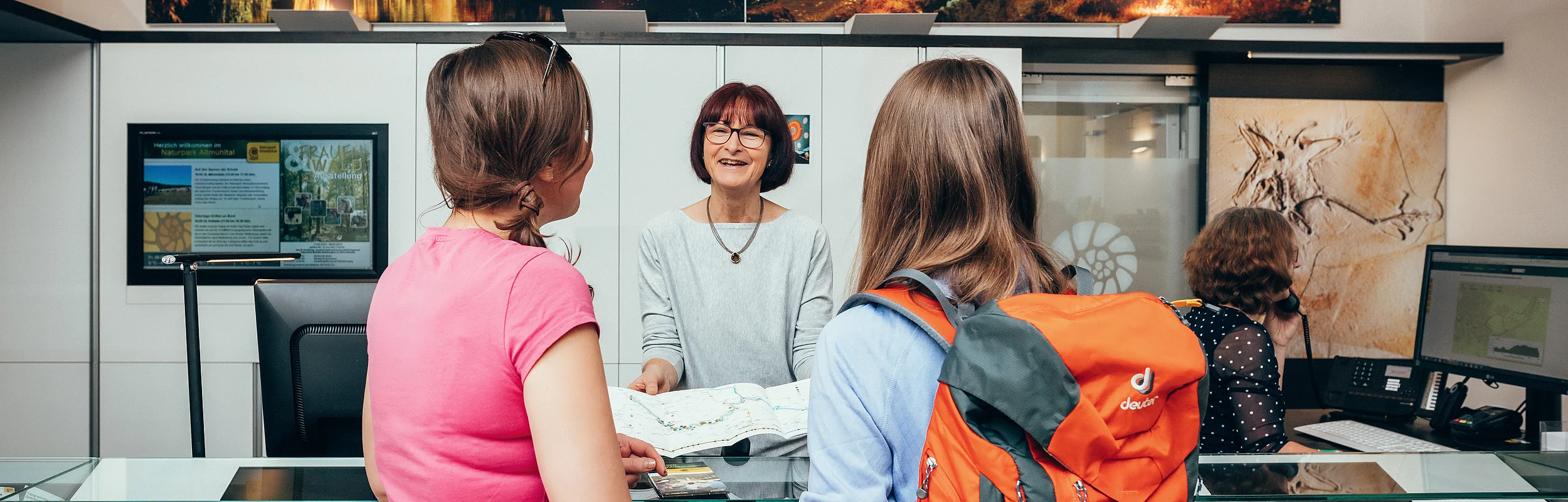 Eine Mitarbeiterin der Tourist-Information berät zwei Kundinnen. Sie hat eine Karte aufgeklappt und erklärt dazu etwas.