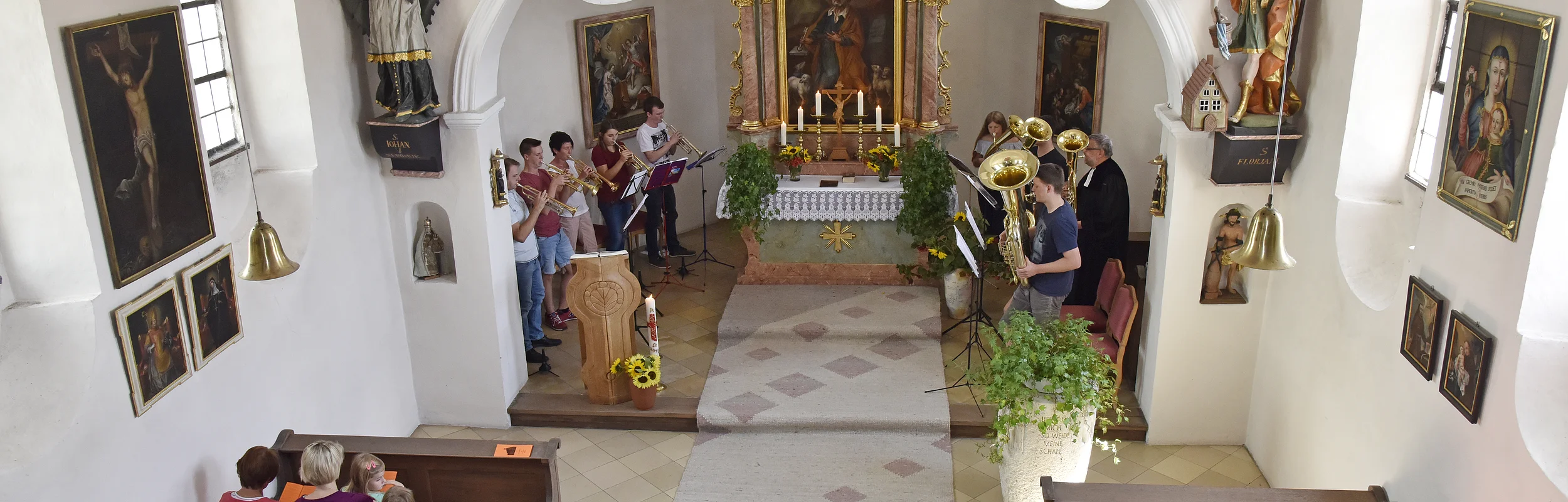 Innenansicht einer Kirche mit Menschen auf Bänken, Musiker spielen vorne Instrumente am Altar.