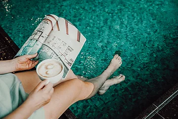 Person hält eine Tasse Kaffee und ein Magazin, Füße im Wasser.