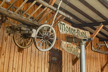 Holzwagenrad und Holzschilder mit der Aufschrift "Motor-Radl Stall" an einer Holzwand.