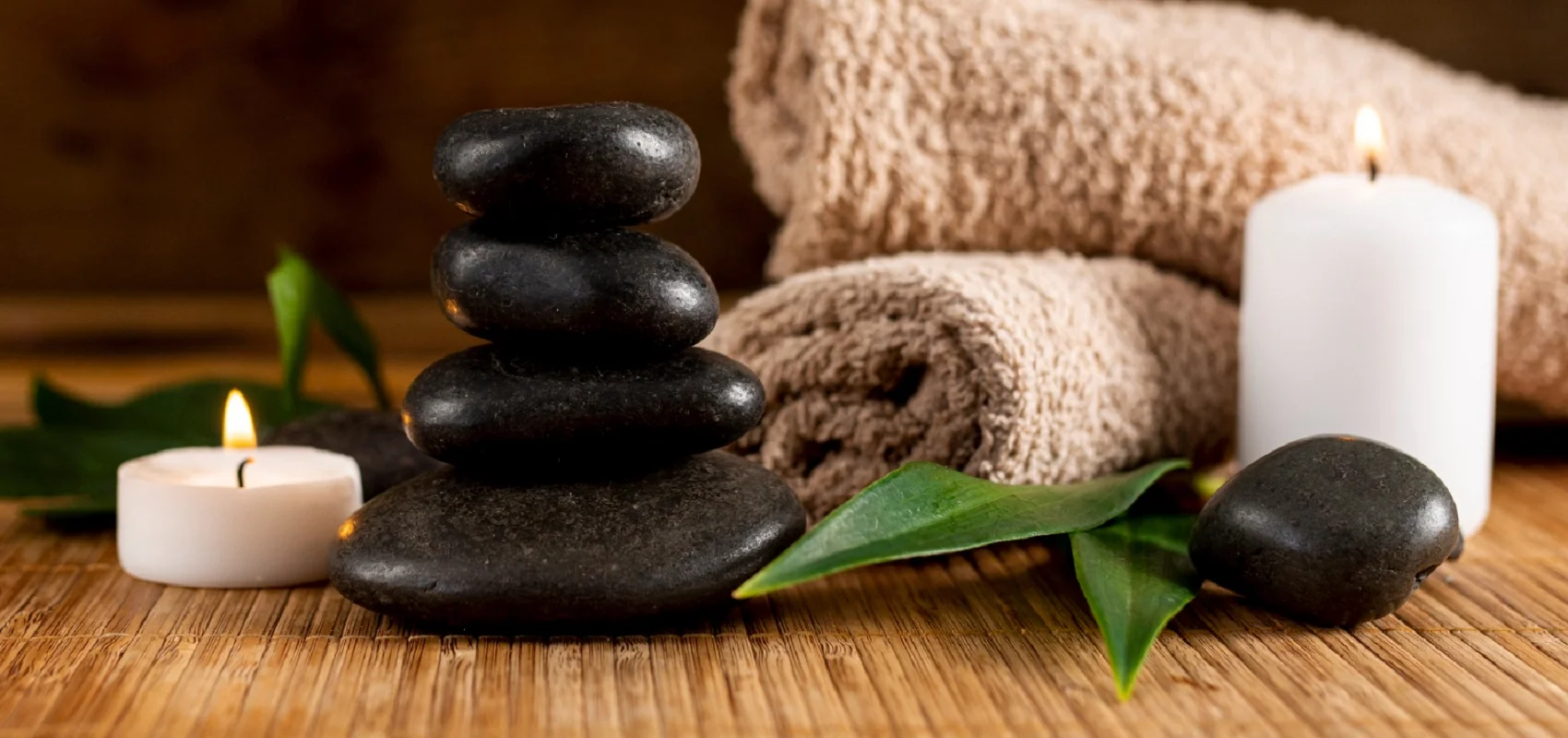 Gestapelte schwarze Steine, brennende Kerzen, ein gerolltes Handtuch und grüne Blätter auf Holzoberfläche.