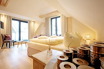 Helles Schlafzimmer mit Doppelbett, großen Fenstern, Holzboden und dekorativen Kissen. Ein Tisch mit Stühlen steht am Fenster.