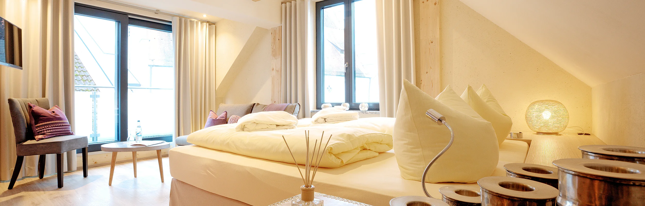 Helles Schlafzimmer mit Doppelbett, großen Fenstern, Holzboden und dekorativen Kissen. Ein Tisch mit Stühlen steht am Fenster.