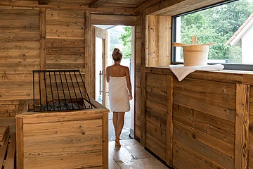 Eine Person in einem Handtuch verlässt eine Holzsauna durch eine Tür. Ein Saunaeimer steht auf einem Handtuch am Fenster.