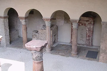 Steinarkaden mit verzierten Säulen in einem historischen Gebäude. Im Hintergrund sind Mauerreste sichtbar.