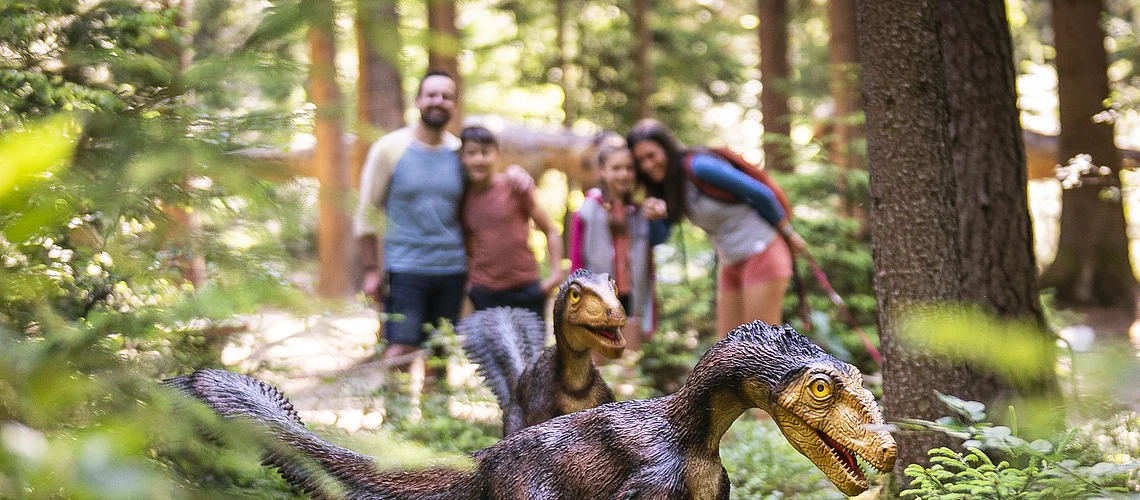 Zwei Dinosauriermodelle im Wald, unscharf im Hintergrund eine Gruppe von Menschen.