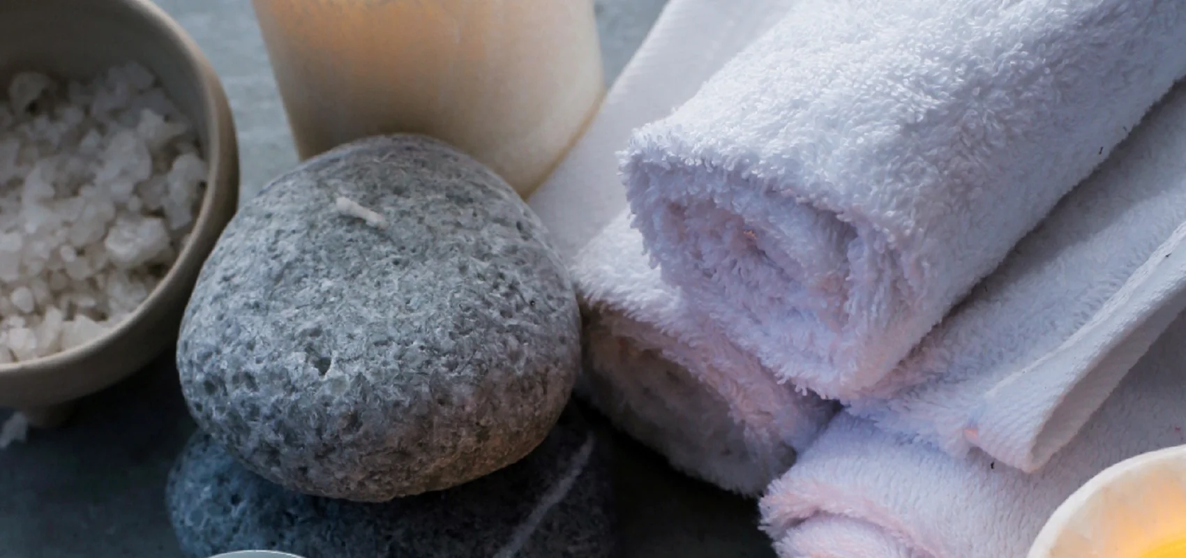 Kerzen, Handtücher, Steine, ein Glas mit Creme, eine Schale mit Salz und ein Holzlöffel auf grauem Hintergrund.