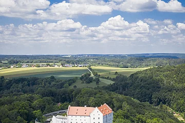 Luftaufnahme eines weißen Schlosses mit rotem Dach, umgeben von Wald und Feldern unter blauem Himmel.