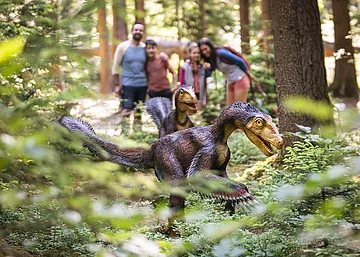 Zwei Dinosauriermodelle im Wald, unscharf im Hintergrund eine Gruppe von Menschen.