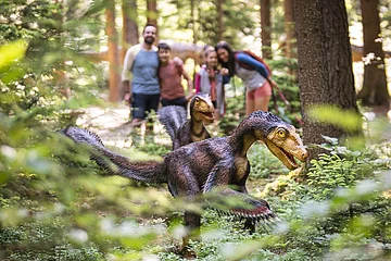 Zwei Dinosauriermodelle im Wald, unscharf im Hintergrund eine Gruppe von Menschen.