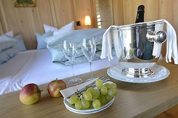Ein Bett mit Kissen im Hintergrund, im Vordergrund ein Tisch mit Sektkühler, Gläsern, Äpfeln und Weintrauben.