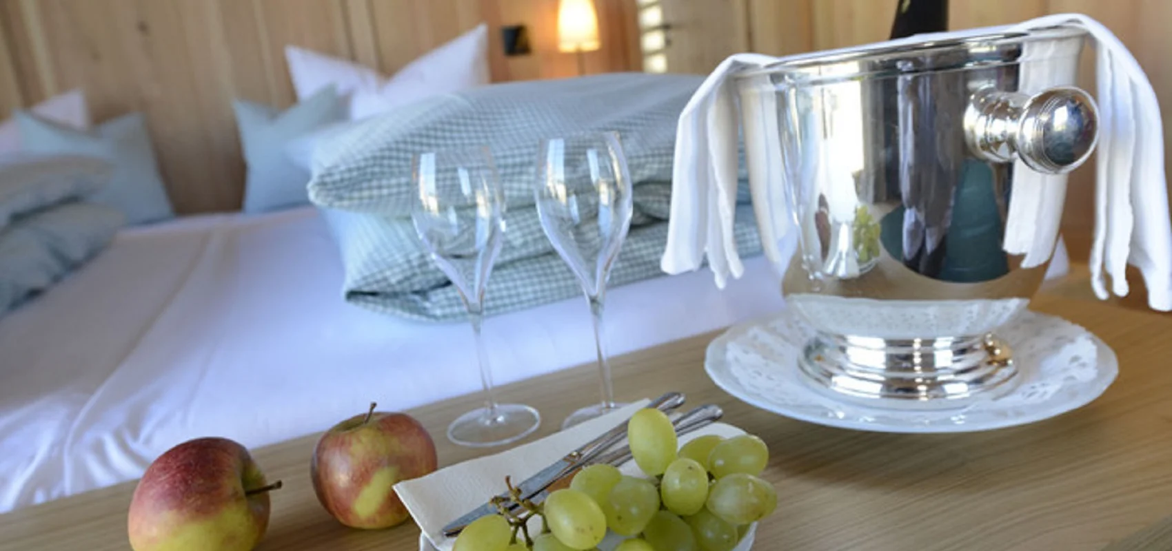 Ein Bett mit Kissen im Hintergrund, im Vordergrund ein Tisch mit Sektkühler, Gläsern, Äpfeln und Weintrauben.