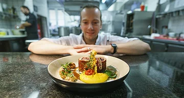 Kulinarisches vom Gasthof Zur Sonne Pappenheim Ein Teller mit kunstvoll angerichtetem Essen auf einer Küchentheke, im Hintergrund ein unscharfer Koch.