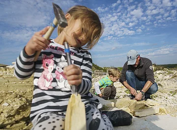 Fossiliensammeln im Steinbruch in Mühlheim Kinder und ein Erwachsener arbeiten mit Hammer und Meißel im Freien auf einem steinigen Gelände.