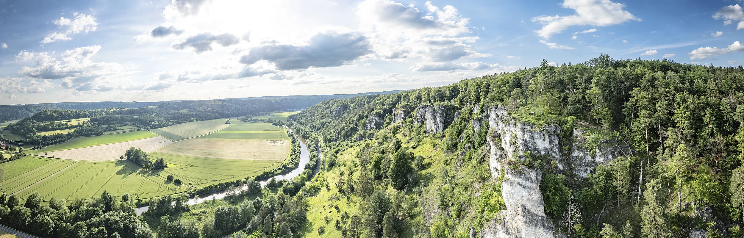 Landschaft mit grünen Feldern, einem Fluss und bewaldeten Hügeln unter einem bewölkten Himmel.