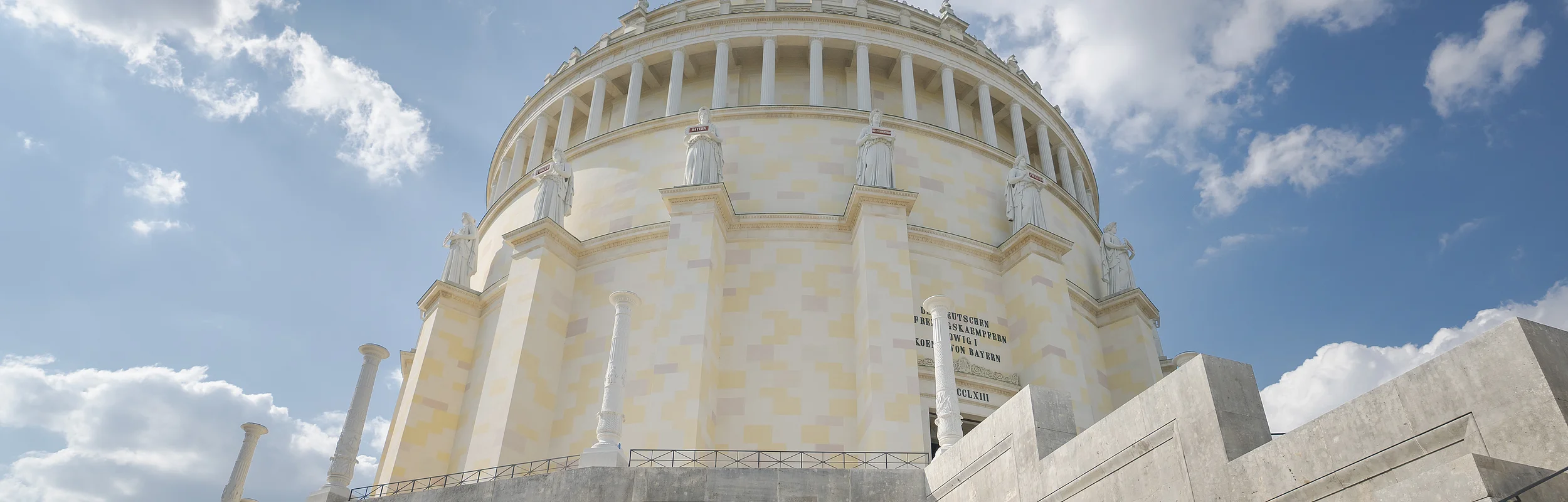 Rundes, klassizistisches Gebäude mit Säulen und Statuen, umgeben von Treppen, unter blauem Himmel mit Wolken.
