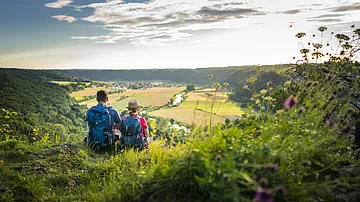 Zwei Personen mit Rucksäcken sitzen auf einem Hügel und blicken auf eine Landschaft mit Feldern und Fluss.