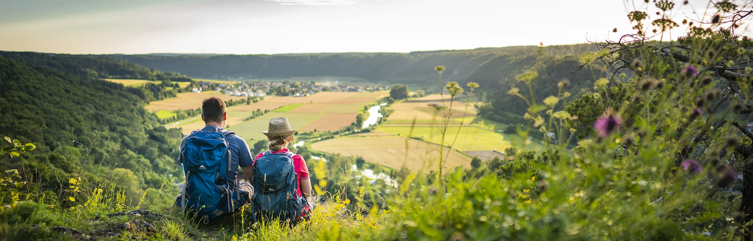 Zwei Personen mit Rucksäcken sitzen auf einem Hügel und blicken auf eine Landschaft mit Feldern und Fluss.