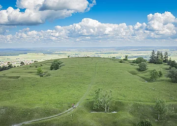Weite grüne Wiesenlandschaft mit vereinzelten Bäumen, im Hintergrund ein Dorf und blauer Himmel mit Wolken.