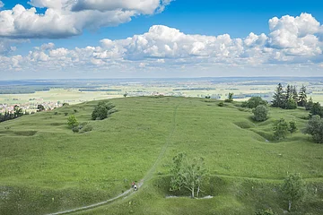 Weite grüne Wiesenlandschaft mit vereinzelten Bäumen, im Hintergrund ein Dorf und blauer Himmel mit Wolken.