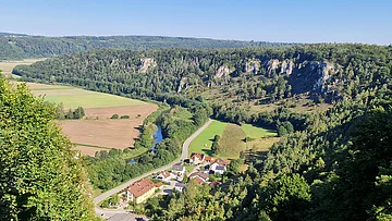 Landschaft mit Fluss, Feldern, Wald und kleinem Dorf im Tal. Hügel im Hintergrund.