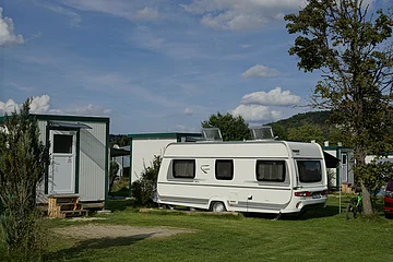 Wohnwagen auf einem Campingplatz neben kleinen Gebäuden und einem Baum.
