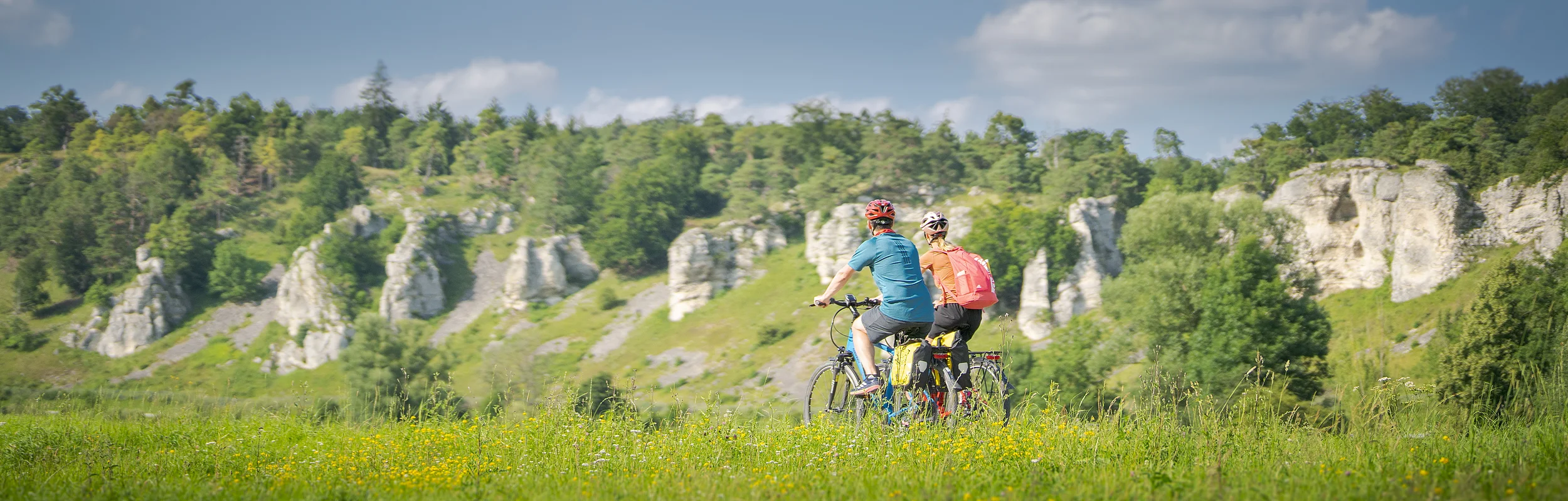 Zwei Personen fahren mit Fahrrädern auf einem grasbewachsenen Feld vor einer Felslandschaft.