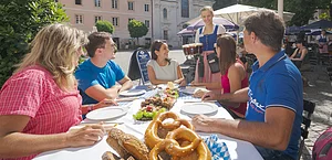 Fünf Personen sitzen an einem Tisch im Freien, eine Kellnerin bringt Bier, Brezeln und Brot liegen auf dem Tisch.