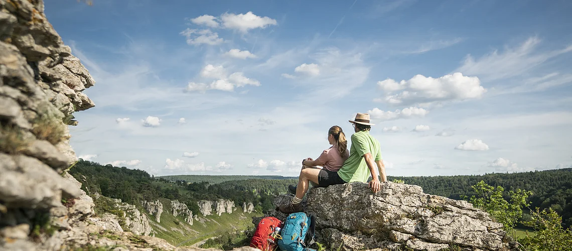Zwei Wanderer sitzen auf einem Felsen und blicken auf bewaldete Hügel unter blauem Himmel mit Wolken.