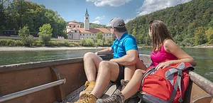Paar sitzt in einem Boot auf einem Fluss und blickt auf ein Gebäude mit Turm und Wald im Hintergrund.