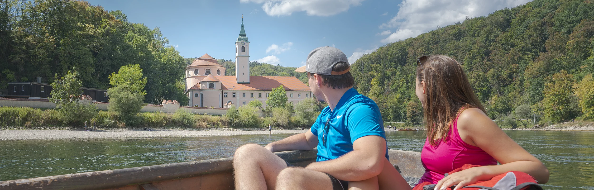 Paar sitzt in einem Boot auf einem Fluss und blickt auf ein Gebäude mit Turm und Wald im Hintergrund.