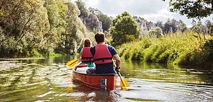 Zwei Personen mit roten Schwimmwesten paddeln in einem roten Kanu auf einem Fluss, umgeben von grüner Natur.
