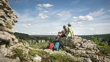 Zwei Wanderer sitzen auf einem Felsen und blicken auf bewaldete Hügel unter blauem Himmel mit Wolken.