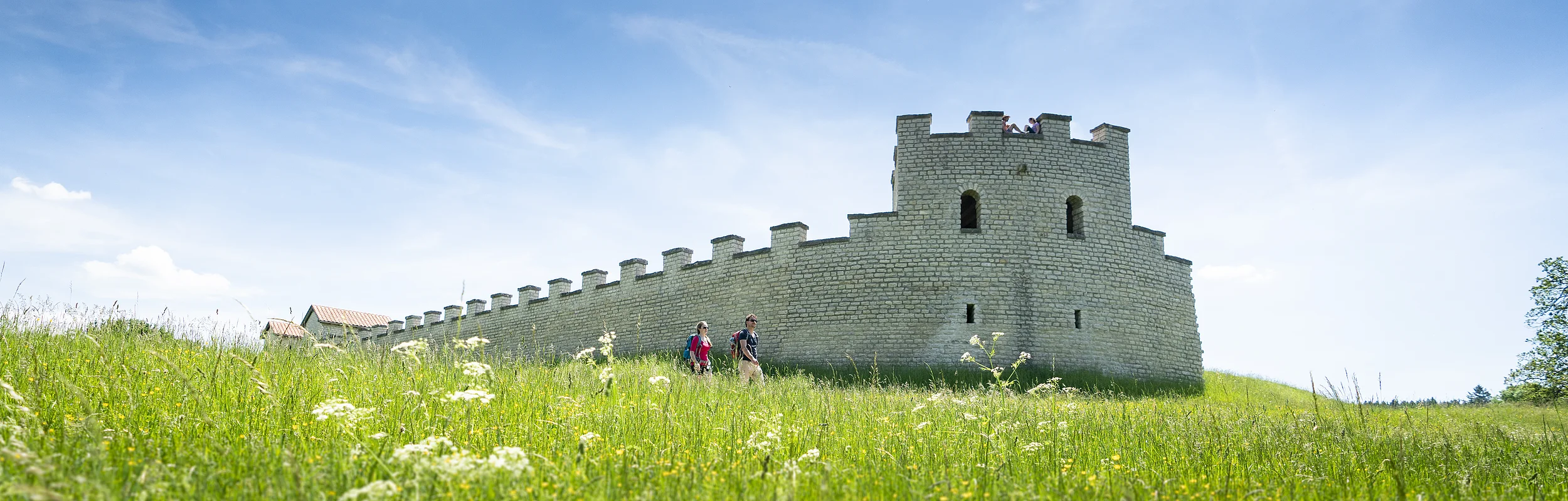 Zwei Wanderer gehen an einer historischen Steinmauer auf einer grünen Wiese unter blauem Himmel vorbei.