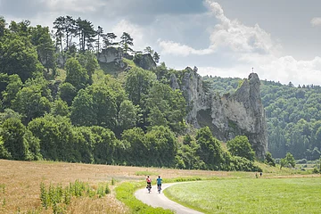 Zwei Radfahrer auf einem Weg vor bewaldeten Felsen mit einem Kreuz auf dem höchsten Felsen.