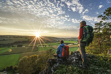 Zwei Wanderer mit Rucksäcken auf Felsen, Blick auf grüne Felder und Sonnenuntergang am Horizont.