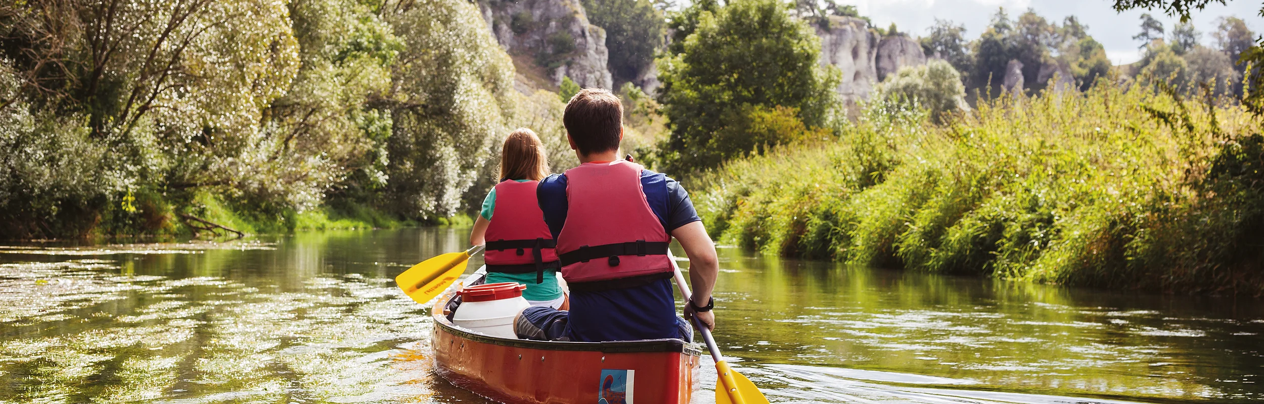 Zwei Personen mit roten Schwimmwesten paddeln in einem roten Kanu auf einem Fluss, umgeben von grüner Natur.