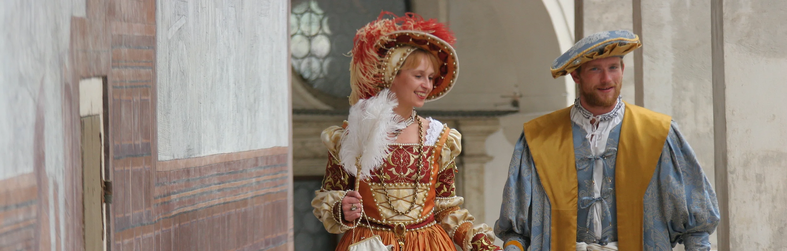 Paar in historischer Renaissancekleidung hält Händchen und geht durch einen Säulengang.