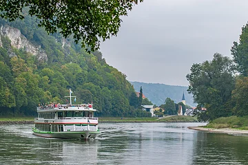 Fahrgastschiff auf Fluss vor bewaldetem Hang und Dorf mit Kirchturm bei bewölktem Himmel