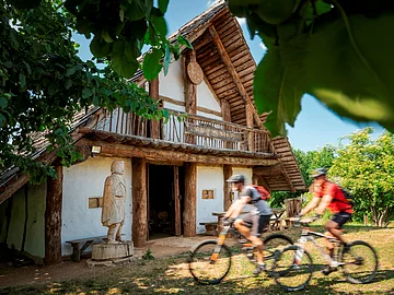 Holzhaus mit Statue davor und zwei Radfahrern, umgeben von Bäumen bei sonnigem Wetter