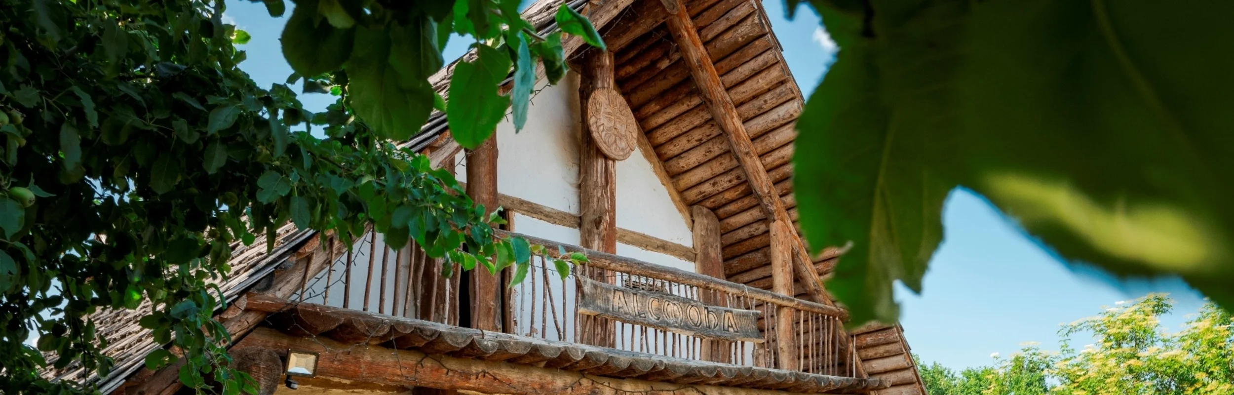 Holzhaus mit Statue davor und zwei Radfahrern, umgeben von Bäumen bei sonnigem Wetter