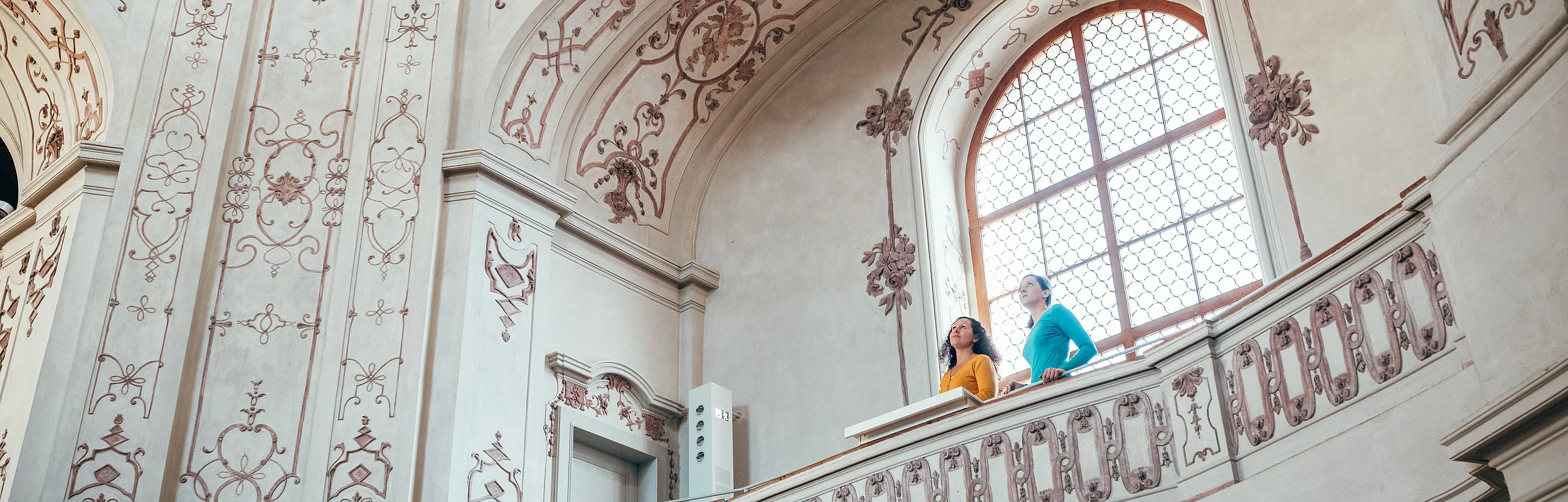 Zwei Personen stehen auf einem verzierten Balkon in einem historischen, mit Fresken geschmückten Innenraum.