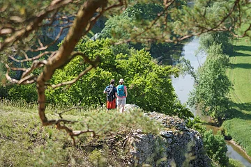 Zwei Wanderer stehen auf einem Felsen mit Blick auf einen Fluss und grüne Landschaft im Tal.