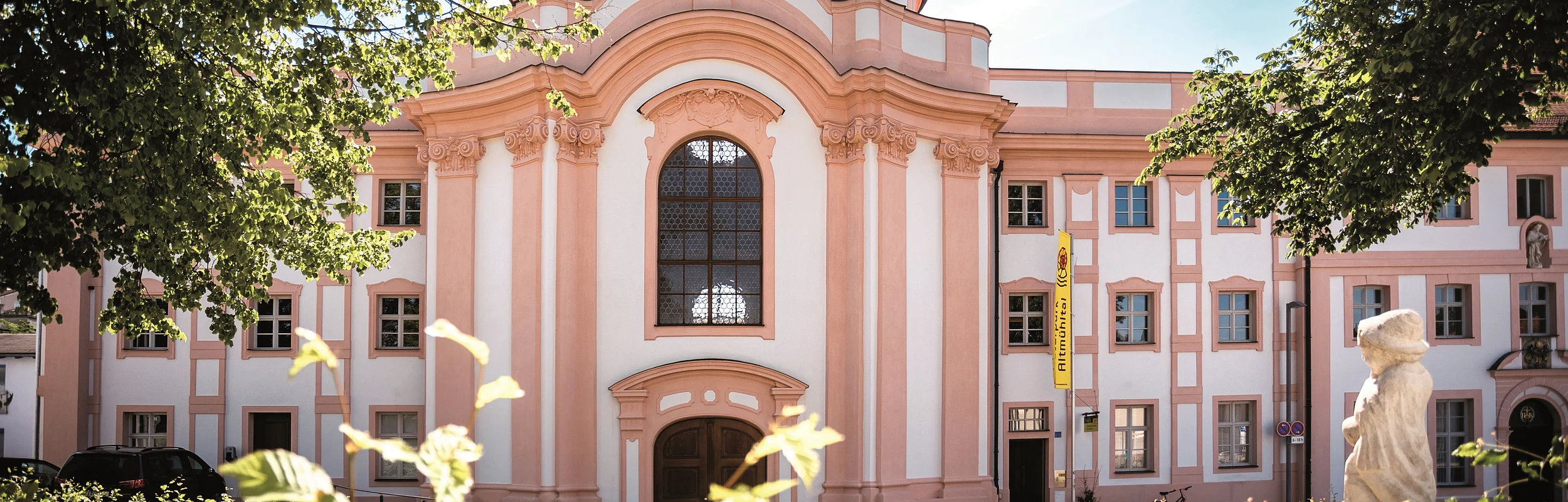 Barocke Kirche mit rosa-weißer Fassade, Statue auf dem Dach und Brunnen mit Skulptur im Vordergrund
